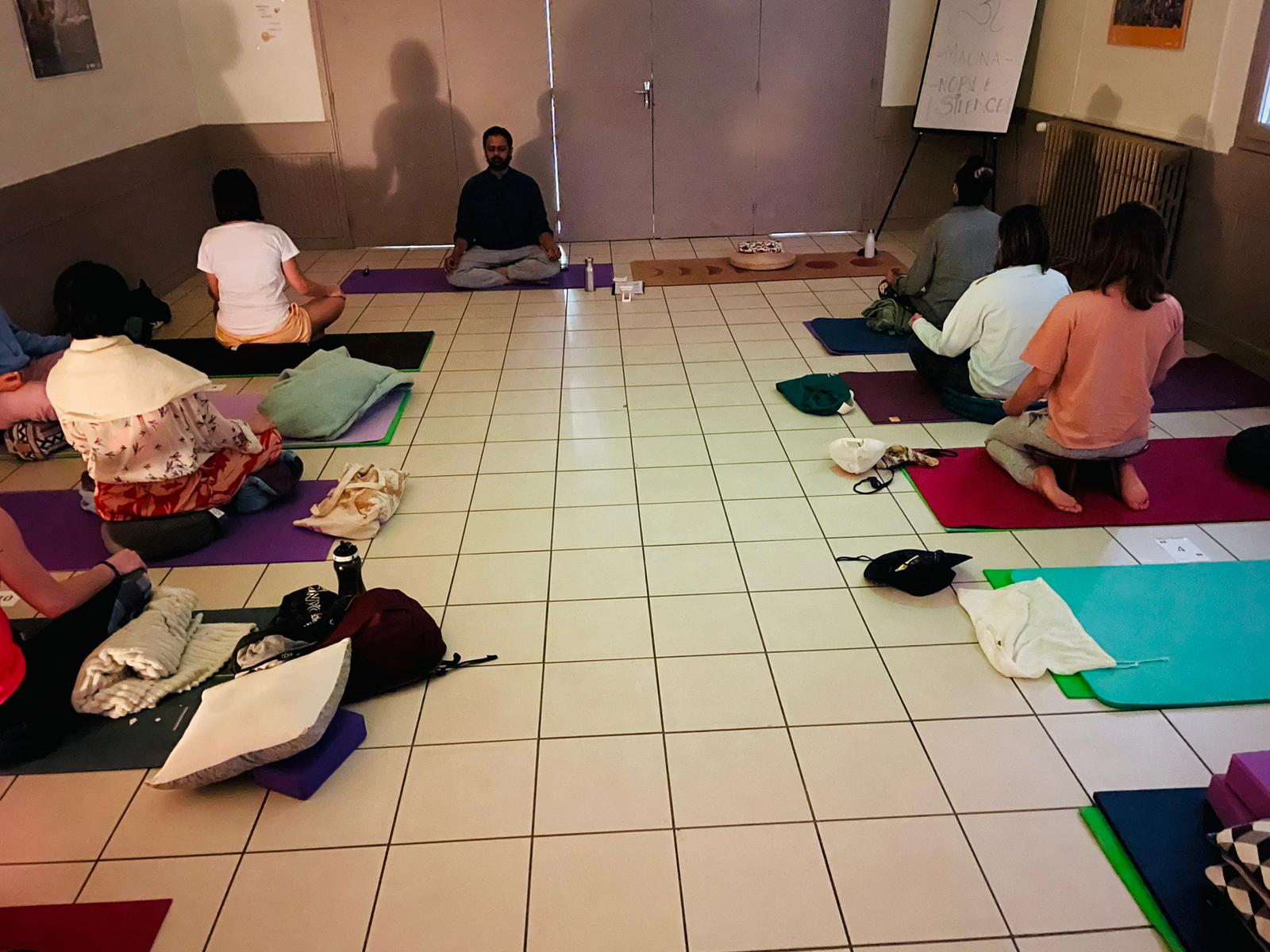 Veer Anand, assis en tailleur au centre de la salle et tourné vers le groupe, guide une méditation ; les pratiquants sont assis en cercle sur leurs tapis autour de lui, lors d'un cours Adinatha à Clermont-Ferrand